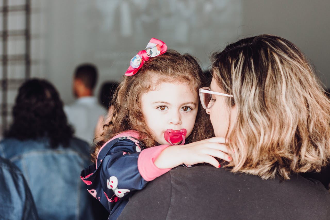 discover A mother lovingly holds her toddler daughter with a pacifier indoors, capturing a moment of care and affection.